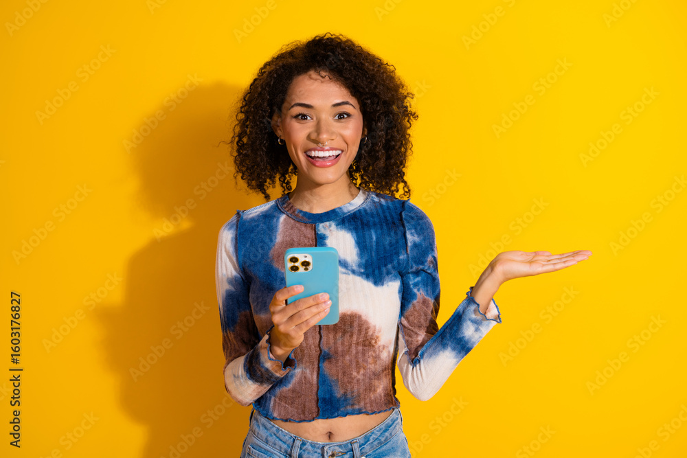 Cheerful young woman with smartphone in colorful outfit gesturing on yellow background expressing joy Cheerful young woman with smartphone in colorful outfit gesturing on yellow background expressing joy