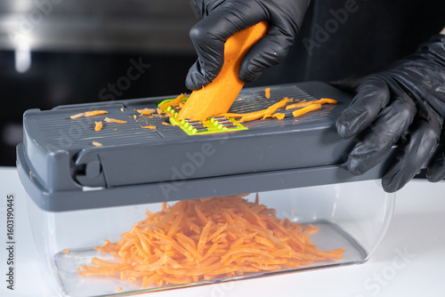 Tableau sur toile Person in black protective gloves expertly grates a fresh orange carrot using a