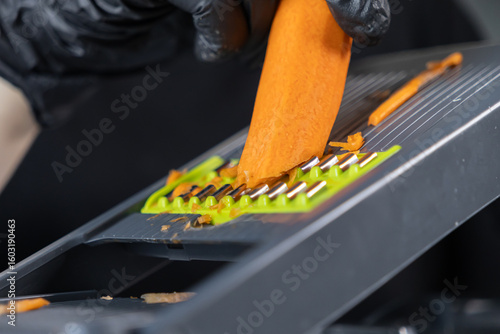 Fotografie Hand in black glove expertly grating a vibrant orange carrot on a modern mandoli