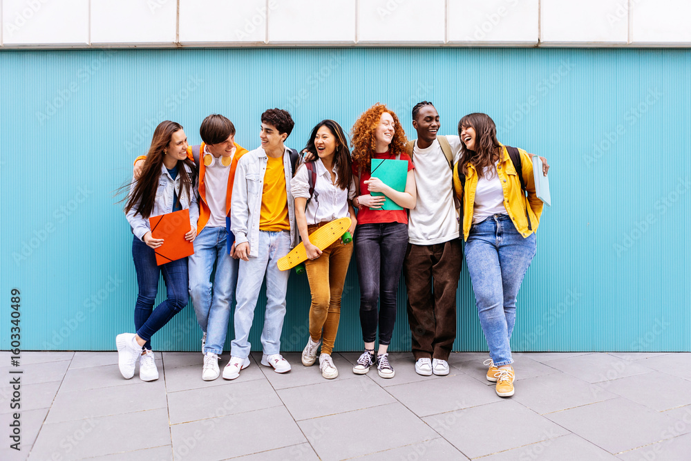 Obraz premium Young happy multiracial student friends standing over isolated blue background. Diverse teenage people having fun talking leaning on campus building college wall. Education and youth concept