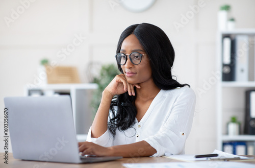 Positive female manager using laptop computer at workplace, typing document, taking part in online business meeting. Young black entrepreneur doing project, participating in webinar