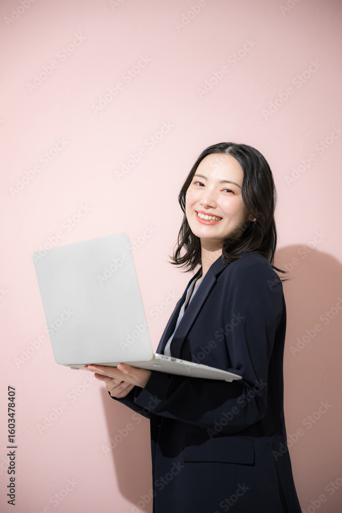 Fototapeta premium パソコン持ってスーツを着た立つカメラ目線の若いかわいい女性のビジネス上半身 ピンク背景の営業などリクルートのイメージ 縦