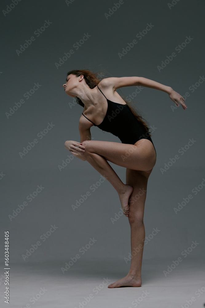 Fototapeta premium graceful woman dancer in black leotard performs contemporary dance pose with extended arms and leg against plain gray background in studio setting