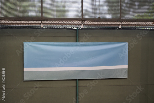 Blank vinyl banner mockup hanging on a green tennis court wall. Empty blue and grey sign for advertising, sponsorship, or text placement.