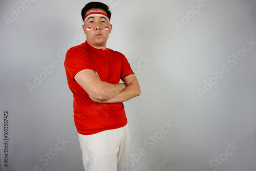 Close-up portrait of a confident young Asian man wearing a red shirt and white pants standing with crossed arms. Indonesian Independence Day concept.