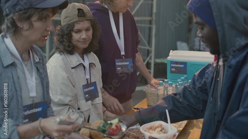 Group of volunteers wearing badges giving hot meals to hungry homeless people at food station during outreach program on urban street