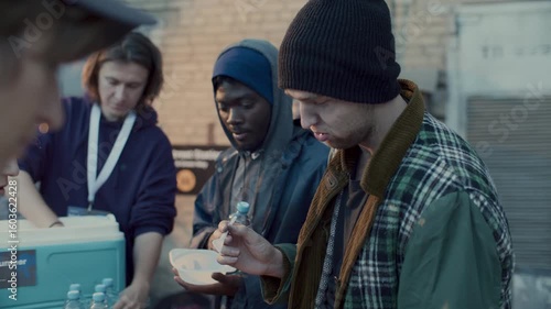 Multi-ethnic homeless people receiving water bottles and bowls of hot meal from volunteers at food drive during outdoor relief event in urban street