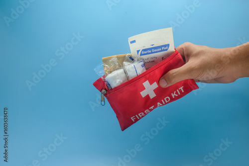 Tableau sur toile A person hand holding a small red first aid kit filled with medical supplies such as gauze pads, tape, and bandages isolated on a blue background
