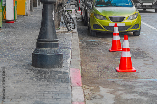 Urban street landscape with sidewalk: empty parking space on side of road with markings, place for car is marked with orange and red plastic traffic cones, there is a bicycle, a lamp post, yellow car