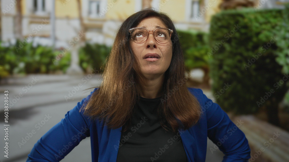 Fototapeta premium Middle-aged hispanic woman holds forehead in pain on sunny urban street; discomfort stress fatigue headache.
