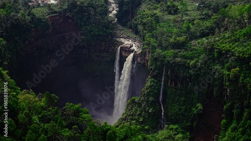 Aerial drone video of hidden waterfall in the tropical forest of East Java, Indonesia, Lush green jungle surrounds the tall waterfall as it flows powerfully down rocky cliffs. 