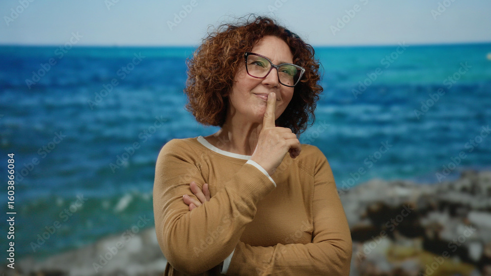 Middle-aged hispanic woman standing outdoors by the seaside with a content expression, wearing glasses and a brown sweater against a backdrop of vibrant blue sea and sky. Middle-aged hispanic woman standing outdoors by the seaside with a content expression, wearing glasses and a brown sweater against a backdrop of vibrant blue sea and sky.