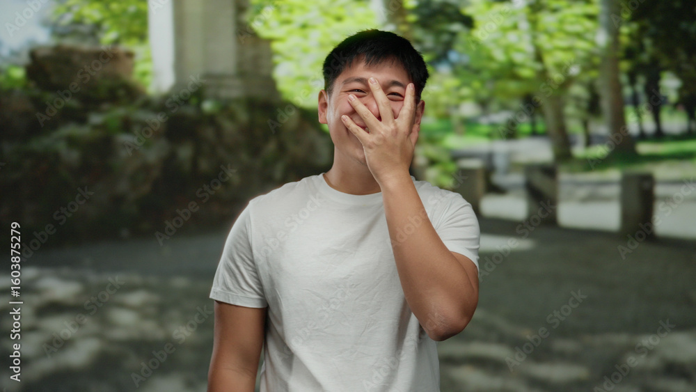 Fototapeta premium Young man laughing and covering his face with his hand outdoors in a vibrant green park setting, conveying a sense of joy and relaxation in nature.