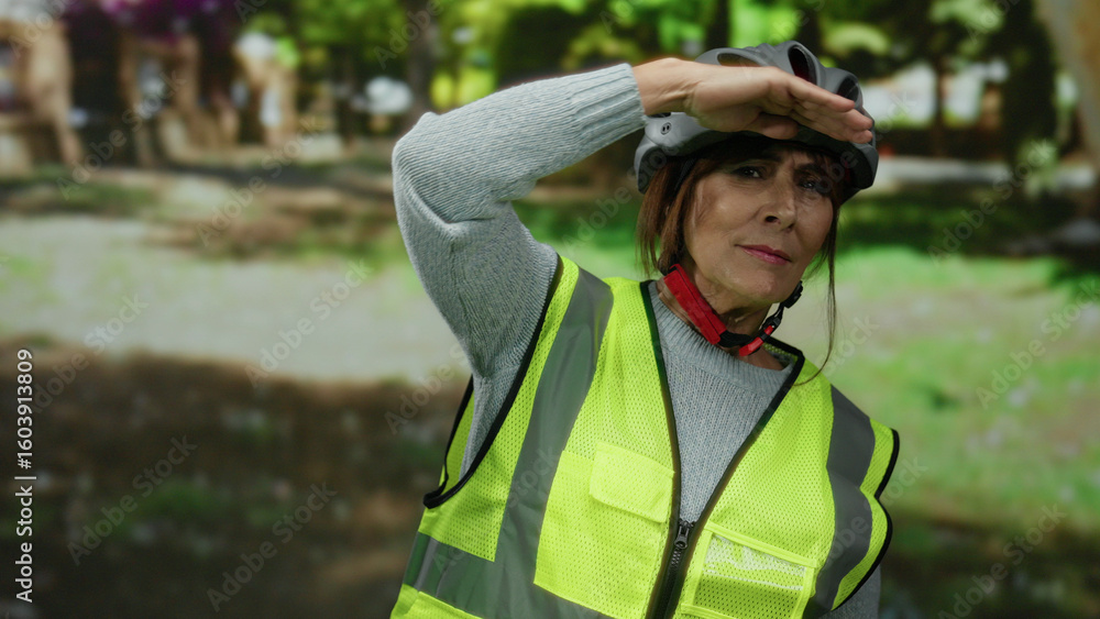 Naklejka premium Senior hispanic woman wearing helmet and reflective vest looks into distance in park setting with trees and greenery surrounding her, embodying outdoor safety and elder activity.