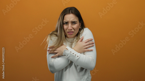 Young blonde woman feeling cold in a white sweater, hugging herself against an isolated orange background, expressing discomfort and shivering slightly.