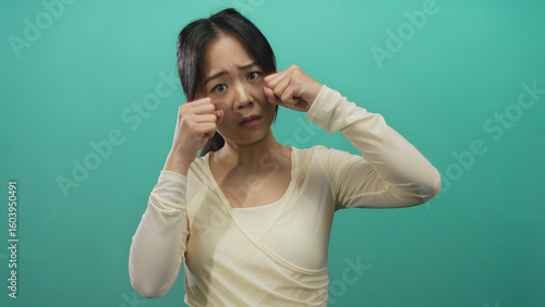 Obraz na plátně Young chinese woman gesturing crying over an isolated green background, expressing emotion in a studio setting with a modern, stylish outfit and thoughtful demeanor