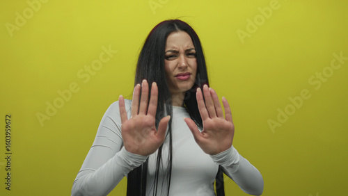 Tablou pe pânză Woman gesturing stop with hands against bright yellow background expressing hesitance and caution in an isolated setting with a strong contrast between her dark hair and attire