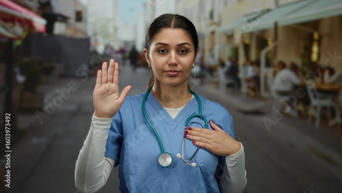 Εκτύπωση καμβά Hispanic woman doctor in blue scrubs makes oath gesture with hand on heart and other raised, standing on a bustling street, embodying professionalism and trust outdoors