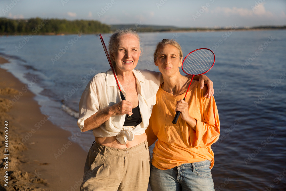 Fototapeta premium portrait Two middle-aged women with badminton rackets stand on the beach and look at the camera on a hot summer evening. Enjoying the little things. spends time in nature in summer. women's friends