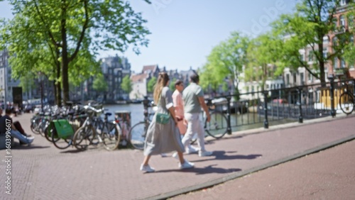 Wallpaper Mural People walking along amsterdam canals with bicycles under sunny skies, featuring blurred urban scenery, pedestrians, trees, and historic buildings in the netherlands. Torontodigital.ca