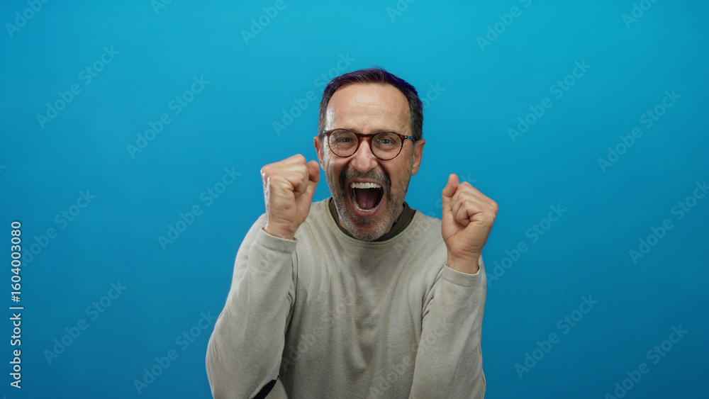 Naklejka premium Mature hispanic man expressing joy and excitement with raised hands against a vibrant blue background, showcasing an enthusiastic and animated expression.