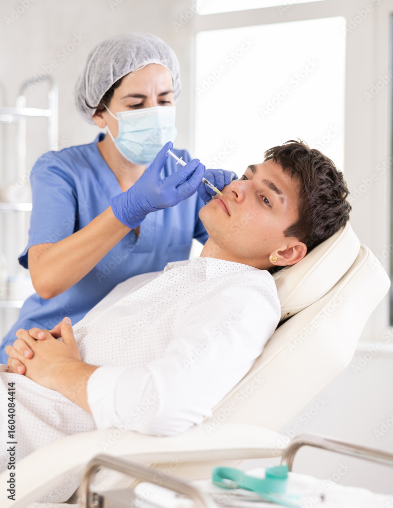 Obraz premium Cosmetologist in medical scrubs, cap and gloves giving filler injection to young man