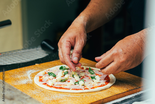 Close-up of hands adding fresh basil leaves to a pizza topped with ham and tomato sauce on raw dough. A step in the homemade pizza-making process captured on a wooden pizza peel.