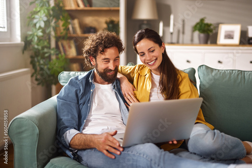 Two people relax on a comfortable sofa, sharing laughs while looking at a laptop screen in a stylish living room filled with natural light and greenery around them.