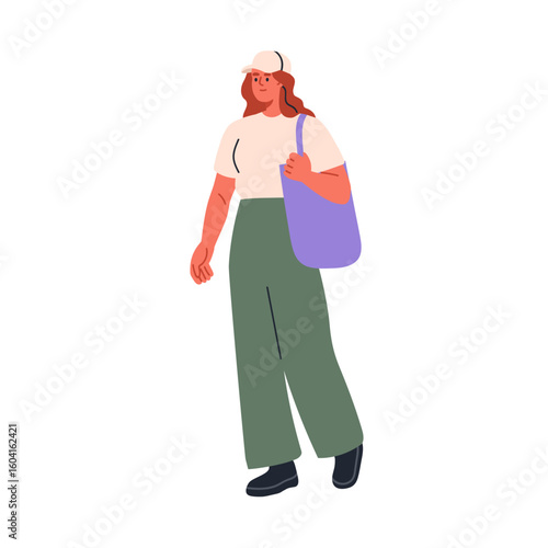 Young woman walking outdoors in casual outfit with baseball cap, carrying tot...
