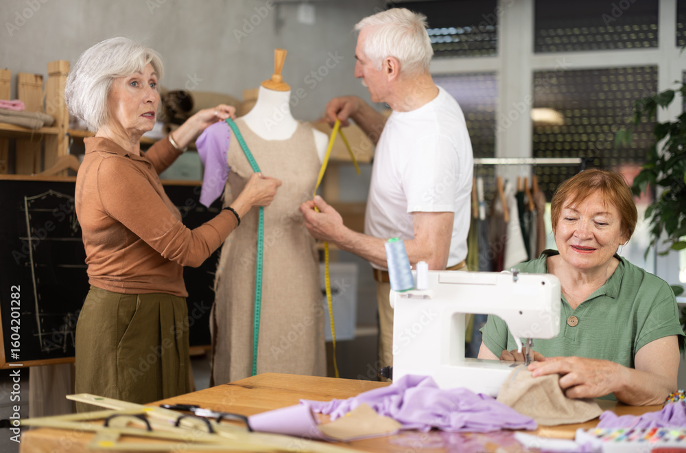Obraz premium Elderly men and women spending time together sewing in cozy workroom, friendly chatting while using machines, cutting fabric and paper templates during hands-on leisure time
