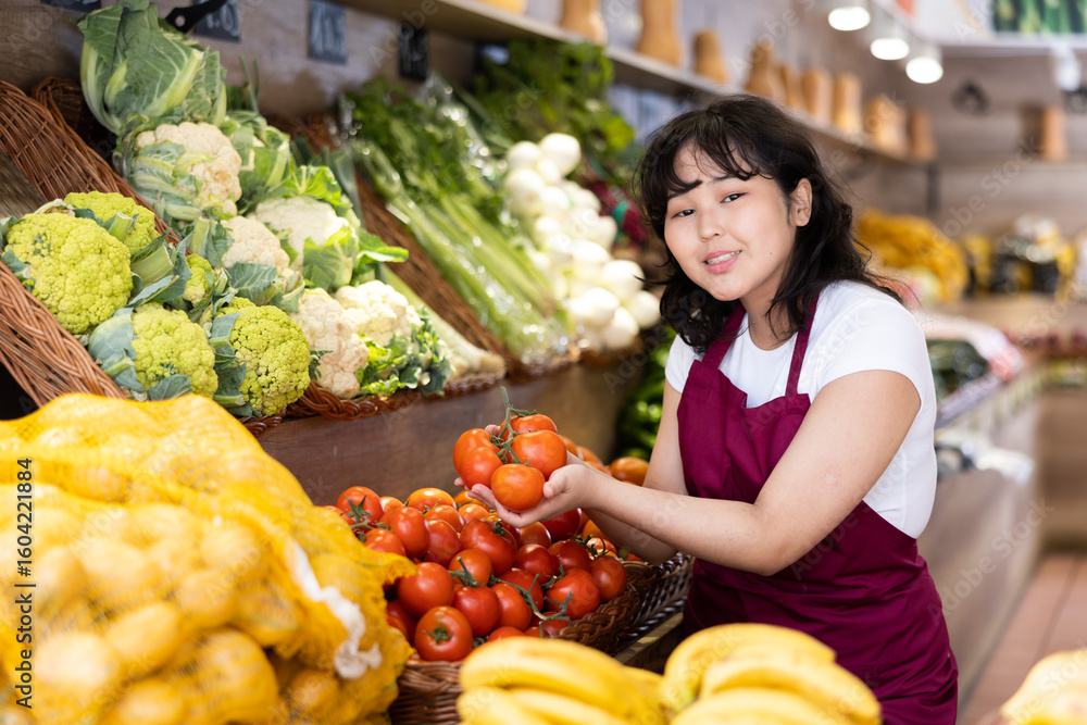 Obraz premium Cheerful friendly young asian salesgirl standing amount produce stands in greengrocery, holding branch of ripe tomatoes while offering fresh organic fruits and vegetables to customers