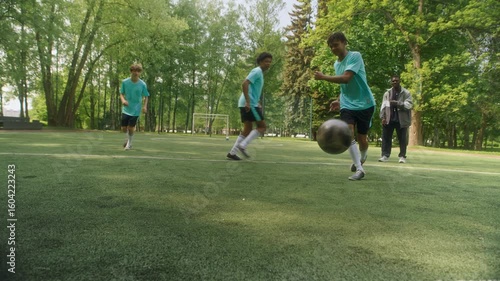 Three young diverse soccer players actively competing on grassy field while one of boys scoring goal toward camera, with Black male coach nearby observing and analyzing action during outdoor training