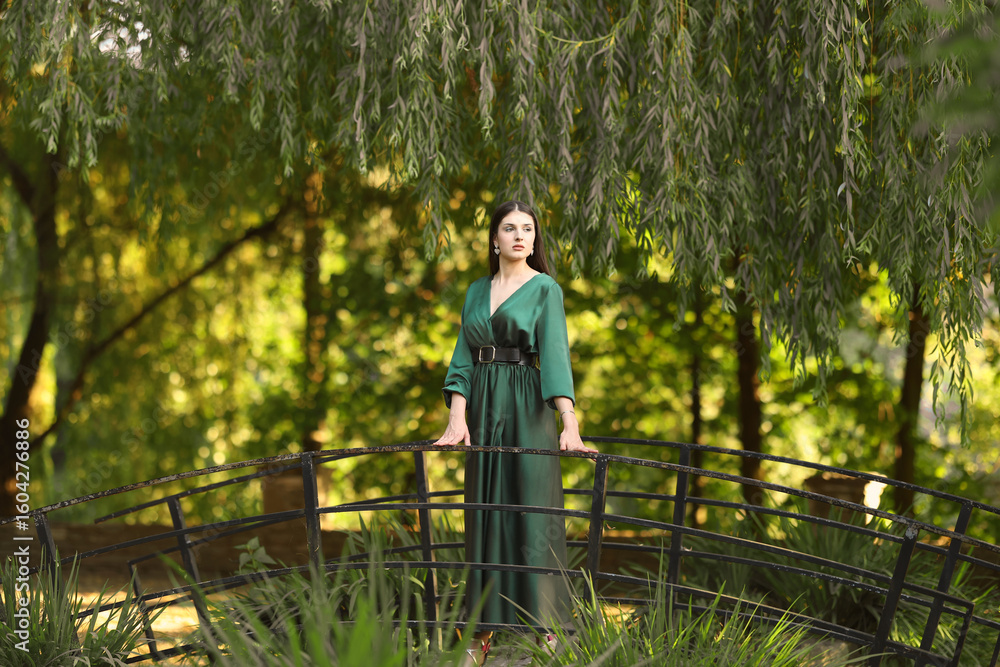 A woman in an emerald green silk dress stands on a small decorative bridge beneath drooping willow branches in soft golden light, exuding serene elegance in nature. A woman in an emerald green silk dress stands on a small decorative bridge beneath drooping willow branches in soft golden light, exuding serene elegance in nature.
