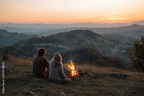 A couple enjoys a campfire on a mountain at sunset, creating a peaceful scene.