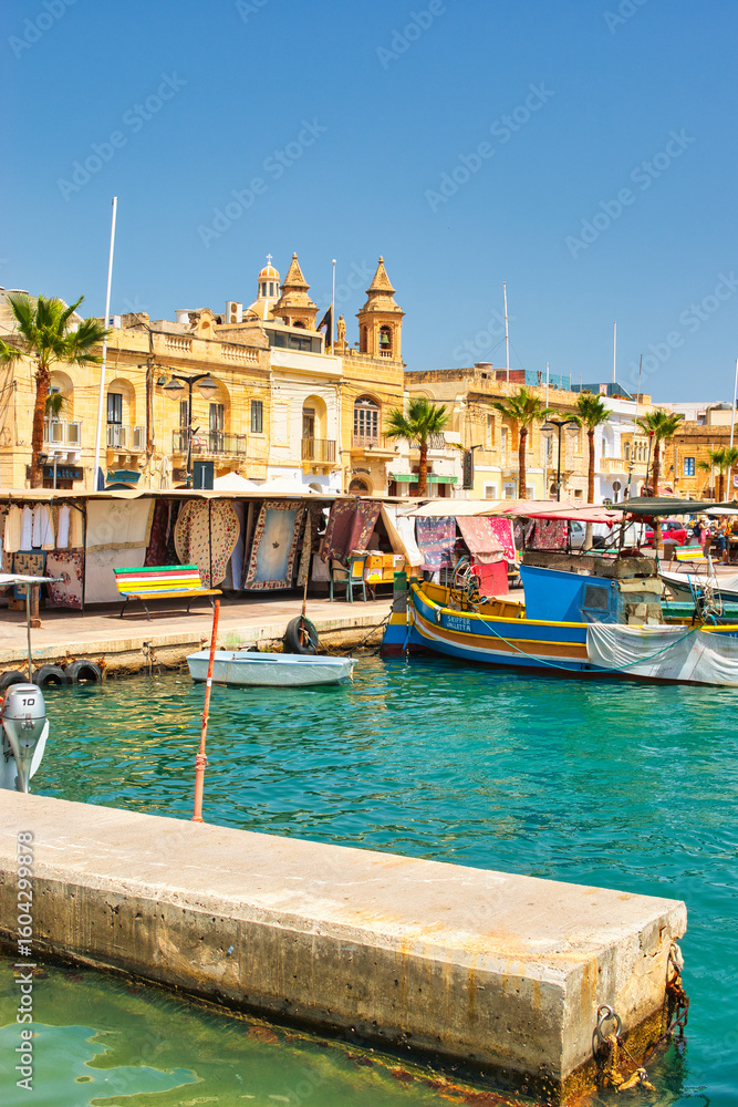 Fototapeta premium Malta Republic, Malta Island. 28. 06.2023. Marsaxlokk city, The harbour and traditional coloured boats on a hot sunny day. This town has a big market every weekend.