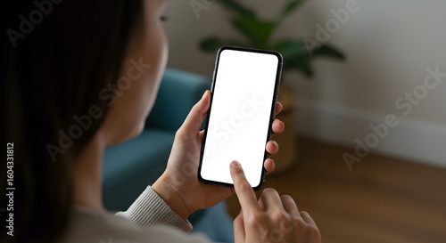 Young woman user holding smart phone in hand touching finger mock up white empty cellphone screen using mobile applications at home. Display mockup close up over shoulder view.