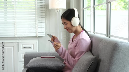 A young Asian housewife relaxes on a sofa, wearing headphones and using her smartphone for online shopping with credit card. She manages household purchases and payments comfortably in modern home.