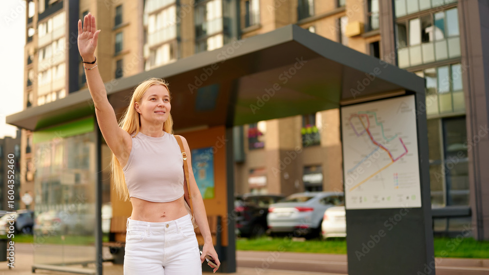 Fototapeta premium woman hailing transportation at bus stop holding smartphone, urban commuter lifestyle. city scene with modern public transport shelter. summertime casual fashion in cityscape.