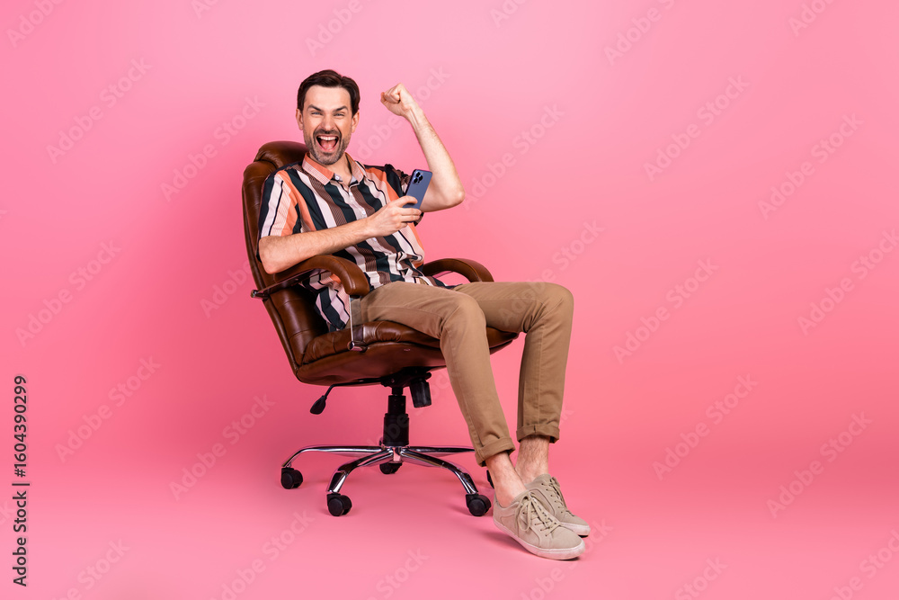 Fototapeta premium Cheerful man seated in chair celebrating success with phone against vibrant pink background in casual fashion