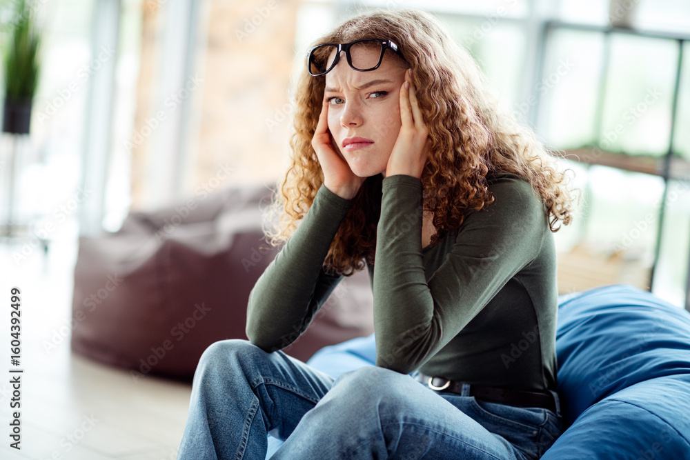 Obraz premium Young woman seated indoors looking thoughtful with glasses on head wearing casual shirt in a modern relaxed interior setting
