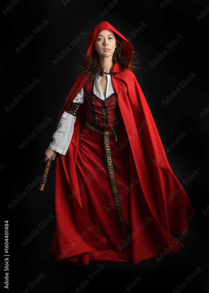dramatic portrait of pretty young female brunette model wearing a red hooded fairytale cloak and medieval historical gown. Holding a axe weapon. Isolated figure on dark studio background. dramatic portrait of pretty young female brunette model wearing a red hooded fairytale cloak and medieval historical gown. Holding a axe weapon. Isolated figure on dark studio background.