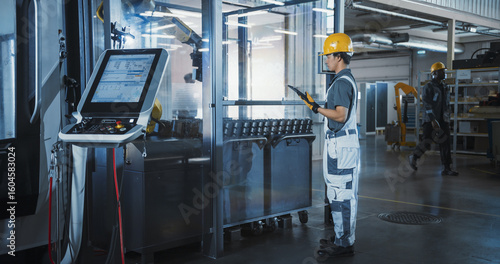 Asian Male Engineer Using Tablet Computer to Control a Robotic Arm at a Metal Components Production Factory. Young Man Working on Manufacturing of Innovative Parts for Artificial Intelligence Systems