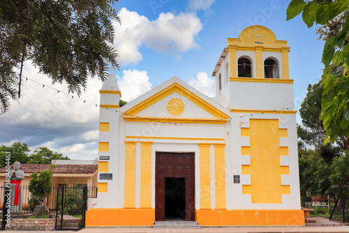 The San Juan Bautista church stands with its characteristic yellow and white facade, dated 1870, in Londres, Catamarca, surrounded by vegetation, under a vibrant sky with clouds.