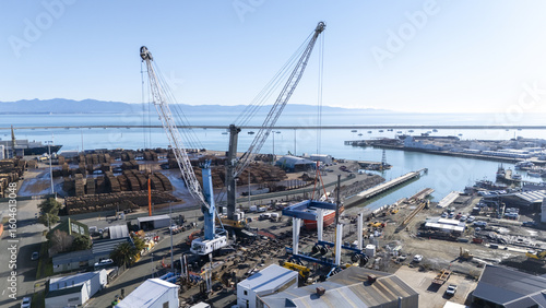 Nelson, New Zealand - 18 June 2025: Aerial view of the port, where towering cranes dominate the landscape, casting long shadows over the bustling docks filled with timber and vessels.