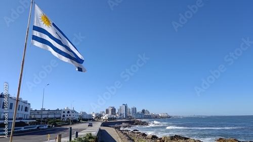 Fotografie The national flag of Uruguay waving in the wind, overlooking a beautiful coastal city with modern buildings and the Atlantic Ocean waves on a rocky shore