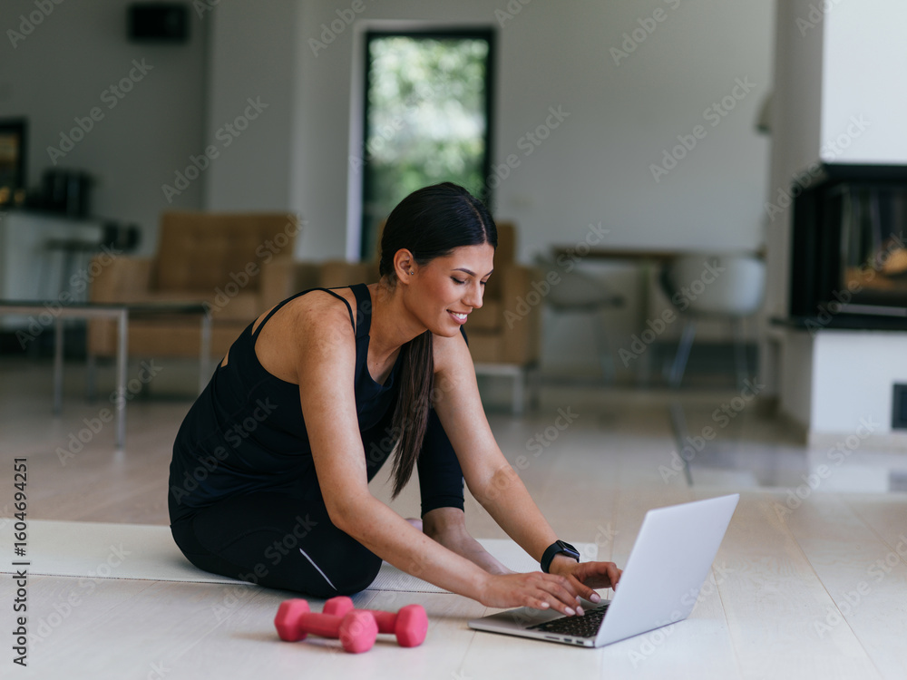 Obraz premium A young woman in sportswear is sitting in the living room and preparing for online training while using a laptop.