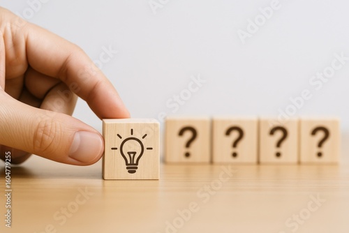 Hand Holding Wooden Block with Light Bulb Icon Next to Four Blocks with Question Marks on a Wooden Surface, Concept of Ideas and Uncertainty