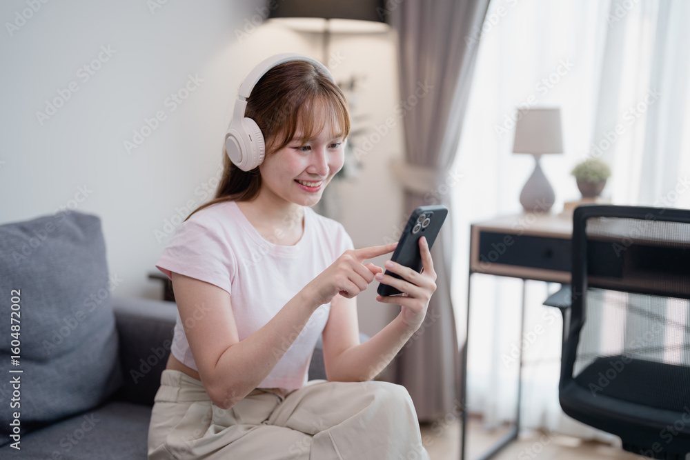 Happy asian woman wearing wireless headphones sitting on comfortable sofa and choosing playlist music from smartphone application in living room, she is relaxing on weekend Happy asian woman wearing wireless headphones sitting on comfortable sofa and choosing playlist music from smartphone application in living room, she is relaxing on weekend