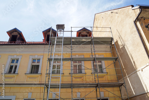 Scaffolding is erected along the facade of a light yellow building with multiple windows and red roof details, indicating construction or renovation work.