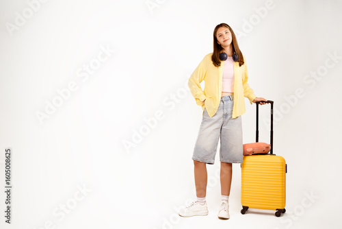 Teenage girl in casual clothes stands with a yellow suitcase and headphones, isolated on a white background. Suitable for travel, vacation, tourism, youth lifestyle, and luggage concepts.